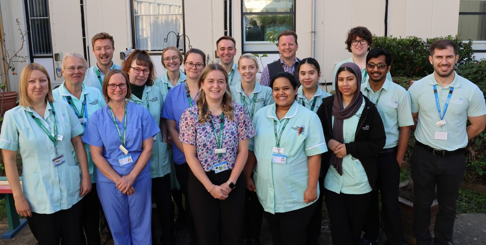 A group of 17 our Pharmacists stand in a group, outside in one of our hospital courtyards, facing the camera and smiling. Many of them wear pale green tunics/ shirts which are the uniform of the pharmacy team and many wear green lanyards. The group are a diverse mix of men and women of different ages and ethnicities.