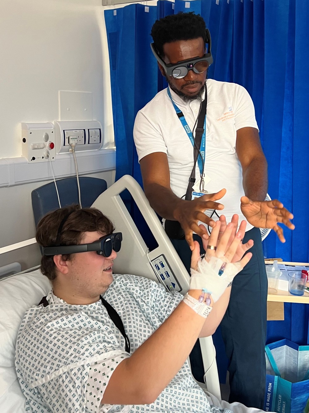 Patient Dylan Turner is lying in a hospital bed, wearing a hospital gown, wearing the goggles which are like thick sunglasses. He has his hands out in front of him, apparently holding an invisible ball, as he gets ready to throw a virtual basketball. Senior Physiotherapist Okechukwu Nwafor stands next to the bed, also wearing goggles and holding his arms outstretched, as though holding the ball.