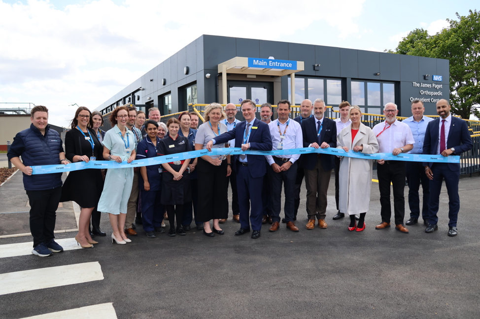 A large group of people are pictured in front of the new building holding a light blue ribbon, which is cut to mark the official opening.